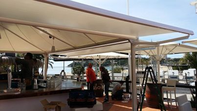 Bar al aire libre con gente, toldo blanco y playa al fondo.