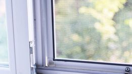 Marco de ventana blanco con mosquitera, ligeramente abierto, que mira hacia un follaje verde borroso.