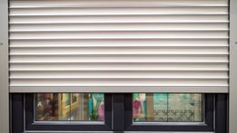 Persiana enrollable de color beige cerrada sobre una ventana, que revela objetos coloridos borrosos en el interior.