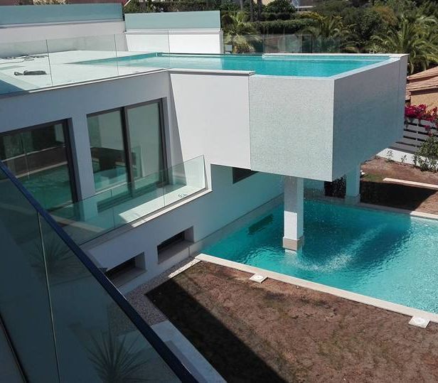 Moderna casa blanca con dos piscinas, una elevada. Agua turquesa, barandillas de cristal y exterior soleado.