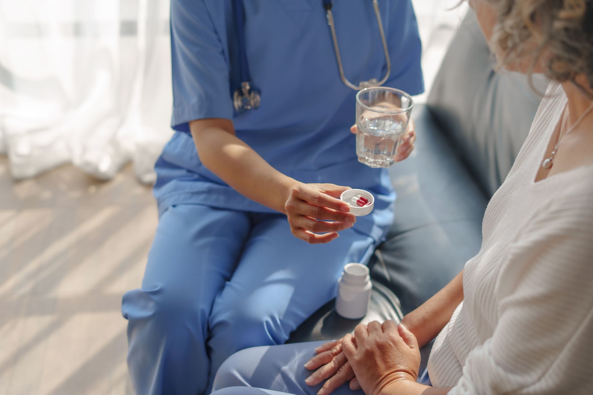 Une aide-soignante en blouse bleue verse de l'eau dans un verre pour un patient assis, qui lui tient la main dans une pièce lumineuse.
