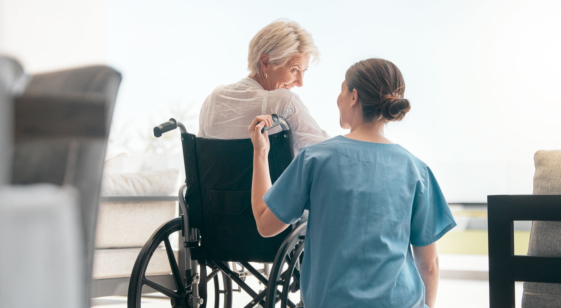 Une aide-soignante en blouse bleue pousse un fauteuil roulant avec une personne âgée dans une pièce lumineuse