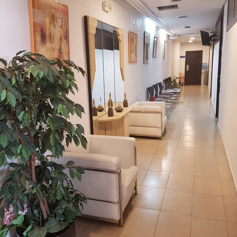 Pasillo con zona de espera: asientos blancos, arte, espejo grande, planta en maceta, azulejos beige y una puerta.