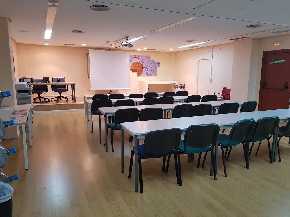 Aula vacía con filas de escritorios y sillas, pantalla de proyector y plataforma elevada con dos escritorios.