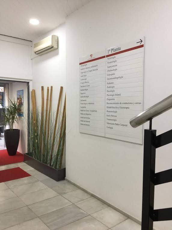 Pasillo con un cartel que indica el primer piso. Escalera con pasamanos, plantas y aire acondicionado en la pared.