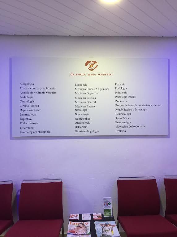 Pared blanca con una lista de servicios médicos y sillas rojas en una sala de espera.