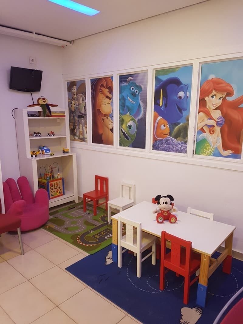 Área de juegos para niños con carteles de personajes de Disney, mesas pequeñas y sillas, además de un asiento rosa con forma de mano.