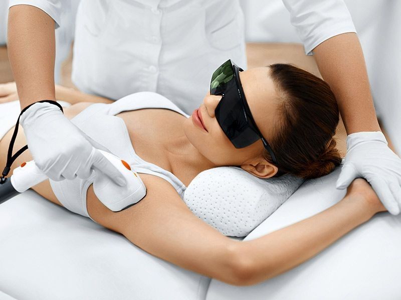 Mujer recibiendo depilación láser en la axila, con gafas y guantes protectores, en un entorno médico.