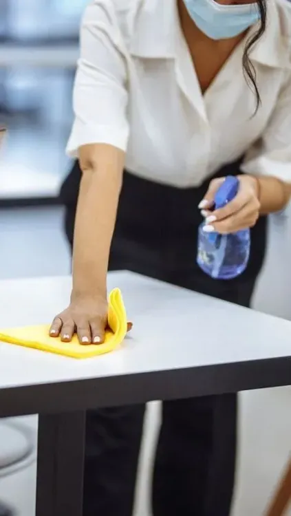 Una mujer que lleva una máscara está limpiando una mesa con un paño y una botella de spray.