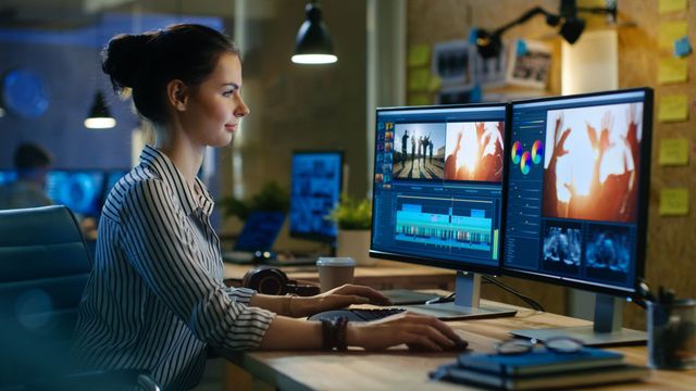 Eine Person sitzt an einem Schreibtisch in einem schwach beleuchteten Büro und bearbeitet Videoinhalte auf zwei Computermonitoren.