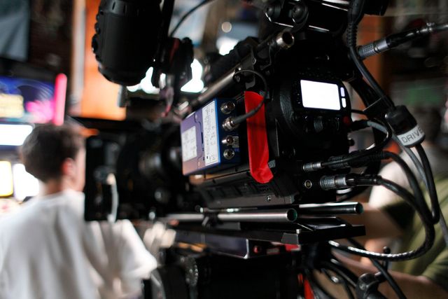 Eine professionelle Kinokamera fokussiert auf eine Szene, während im Hintergrund eine Person am Filmset verschwommen zu sehen ist.