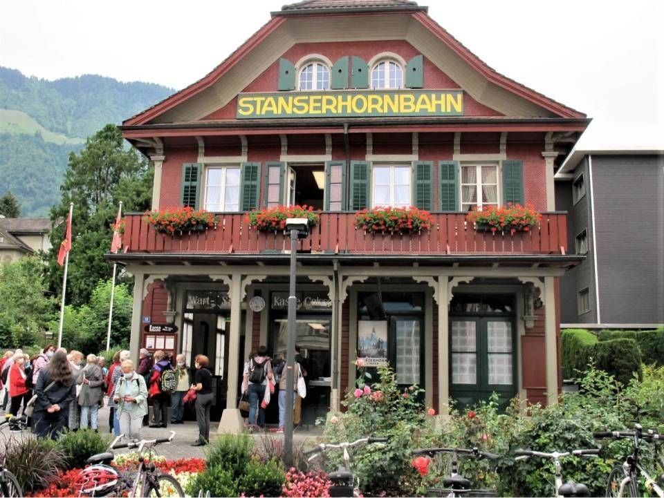 Das Bahnhofsgebäude Stanserhorn in der Schweiz, vor dem sich Menschen versammelt haben, umgeben von Blumen und Bäumen.