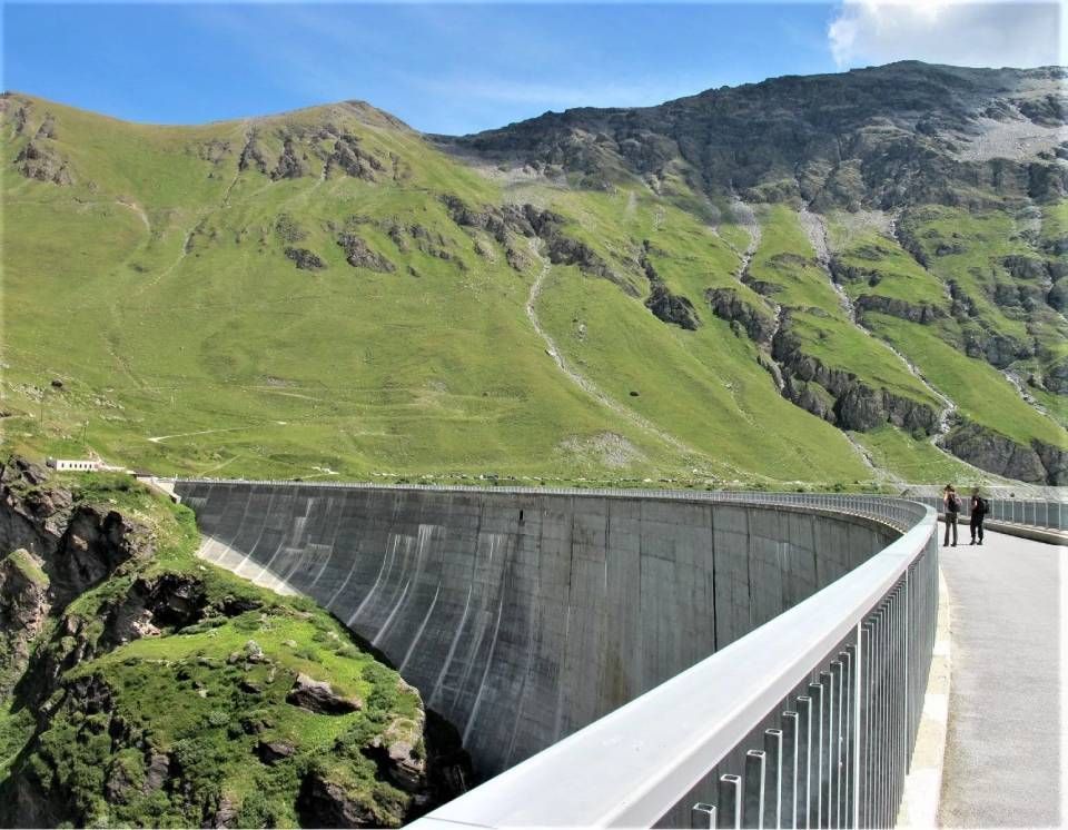 Eine geschwungene Staumauer aus Beton in einer Berglandschaft, betrachtet von einem Steg mit Metallgeländer.