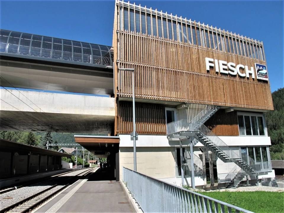 Der moderne Bahnhof Fiesch mit seiner holzlattenverkleideten Fassade und der Metalltreppe, gesehen vom Bahnsteig an einem sonnigen Tag.