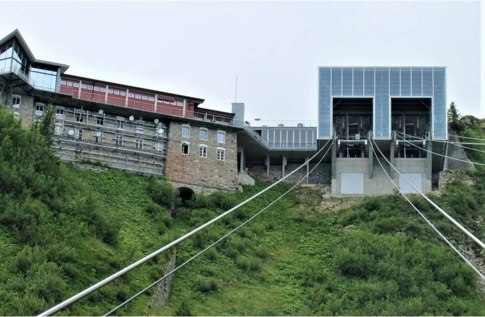 Ein mehrstöckiges Gebäude aus Stein und Glas, das an einem grasbewachsenen Berghang thront, von dessen unterer Fassade dicke Kabel herabhängen.