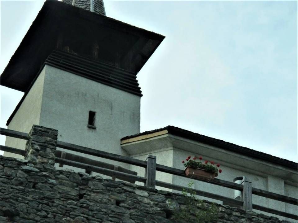 Über einer Steinmauer mit einem Holzzaun und einem Blumenkasten erhebt sich ein steinerner Kirchturm mit einem weit überhängenden Dach.