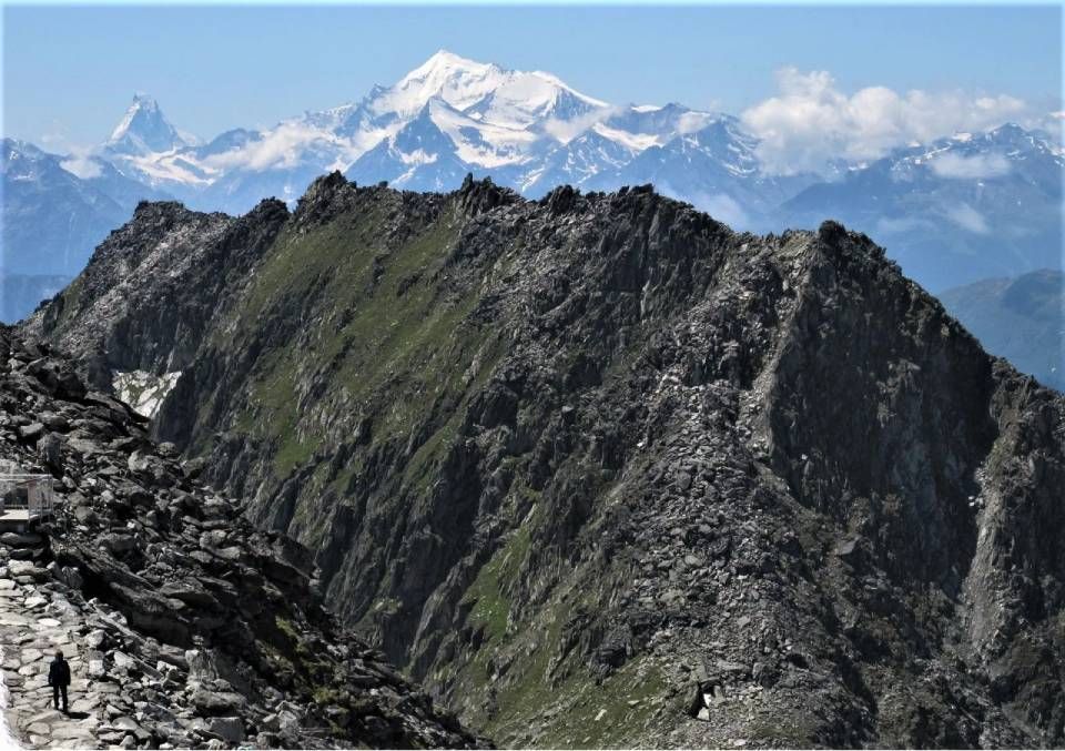 Ein einsamer Wanderer geht einen felsigen Bergpfad entlang, im Hintergrund ein zerklüfteter Grat und das schneebedeckte Mont-Blanc-Massiv.