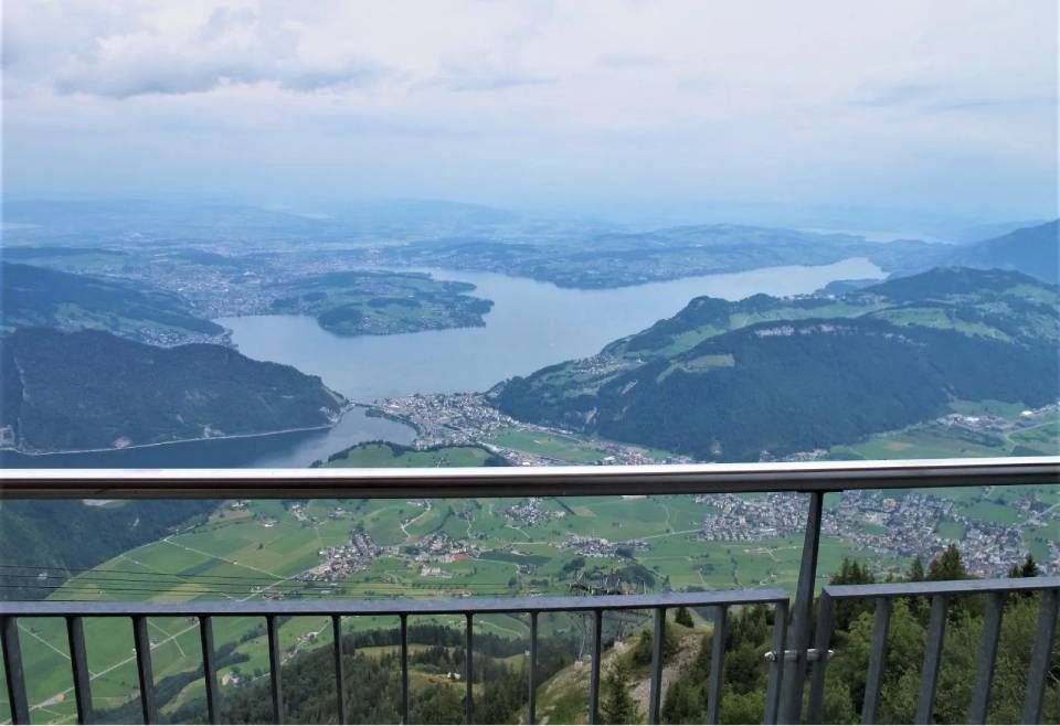 Ein Blick aus der Vogelperspektive auf einen See, eingebettet zwischen sanften grünen Bergen, und eine Stadt, die hinter einem Metallgeländer zu sehen ist.