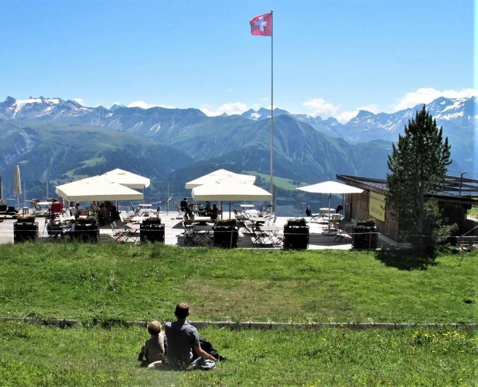 Eine Person und ein Kind sitzen unter einer Schweizer Flagge auf einem grasbewachsenen Hügel mit Blick auf die Terrasse eines Cafés und eine Bergkette.