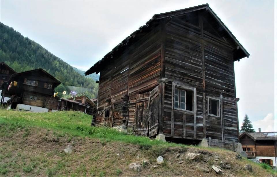 Eine alte, dunkle Holzscheune steht an einem grasbewachsenen Hang in einer Bergregion; in der Nähe sind weitere rustikale Gebäude zu sehen.