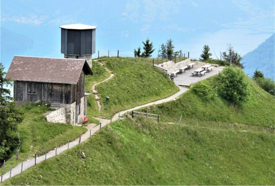 Auf einem üppig grünen Grashügel mit malerischem Pfad befinden sich ein kleines, rustikales Holzgebäude und ein moderner Aussichtsturm.
