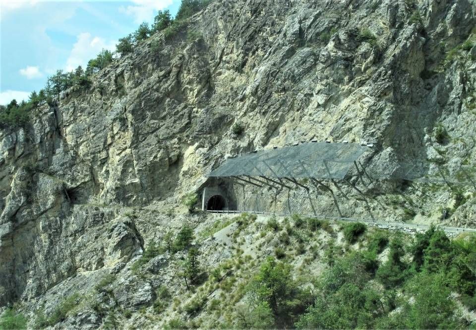 Ein in eine Felswand gehauener Bergtunneleingang, geschützt durch einen grossen, schrägen Steinschlagschutz aus Metall.
