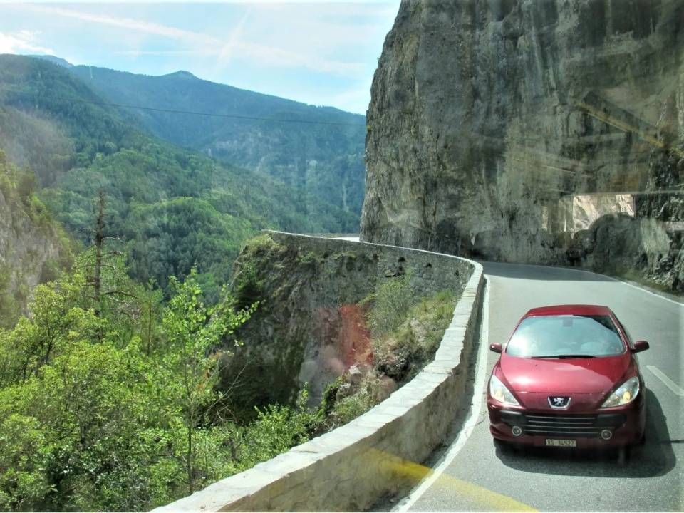 Ein roter Peugeot fährt auf einer schmalen, an einer Klippe entlangführenden Strasse mit Blick auf ein üppiges, gebirgiges Tal.