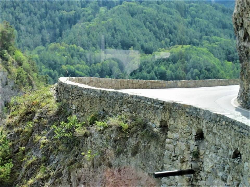 Eine geschwungene Strasse mit einer steinernen Stützmauer grenzt an eine steile Klippe, die einen dichten grünen Wald überblickt.