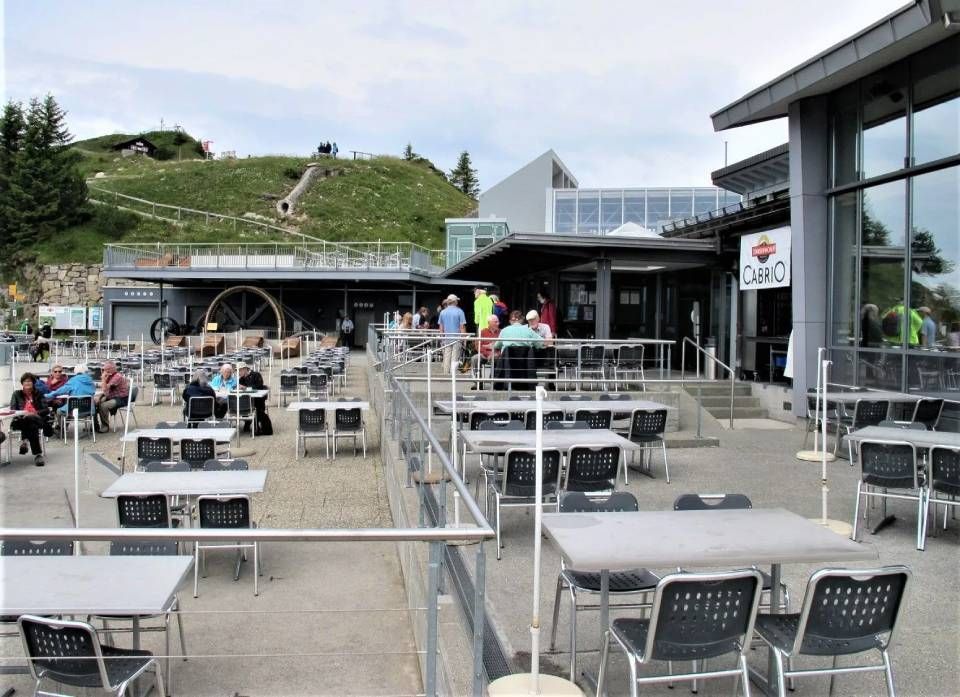 Sitzbereich auf einer Terrasse mit Tischen und Stühlen, angrenzend an ein Gebäude mit einem „Cabrio“-Schild, in der Nähe einer hügeligen Landschaft.