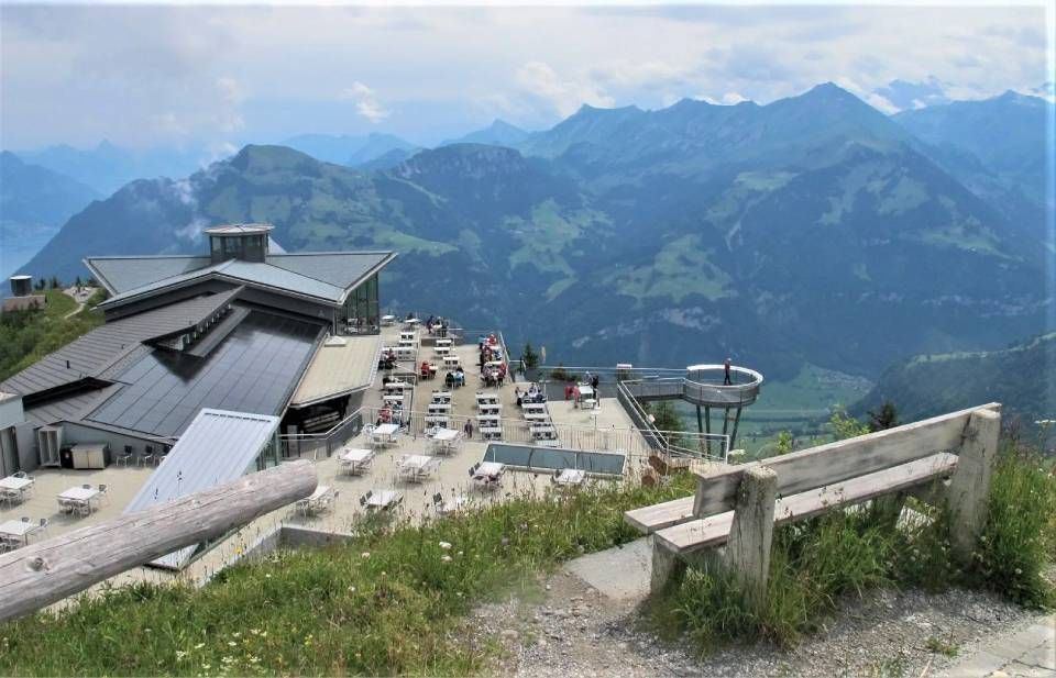 Ein erhöhter Blick auf die Terrasse eines Bergrestaurants mit Sitzgelegenheiten im Freien, einer modernen Aussichtsplattform und alpiner Landschaft.