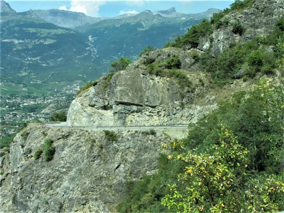 Eine schmale Asphaltstrasse schlängelt sich an einer zerklüfteten, felsigen Bergklippe entlang, im Hintergrund ist ein Dorf im Tal zu sehen.