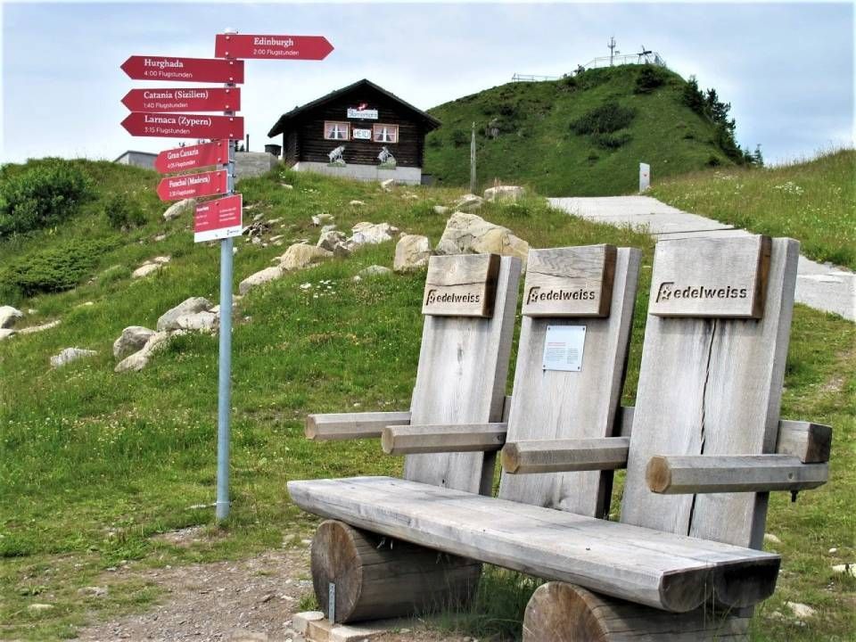 Auf einem grasbewachsenen Hügel steht eine Holzbank mit drei Sitzen neben einem Wegweiser mit mehreren roten Schildern.