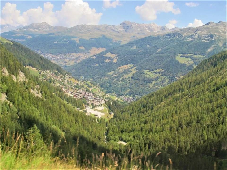 Ein kleines Alpendorf, eingebettet in ein üppig grünes Bergtal, umgeben von bewaldeten Hängen und fernen Gipfeln.