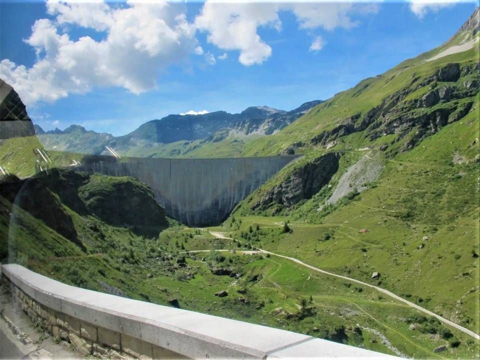 Ein Betonstaudamm eingebettet in ein üppig grünes Bergtal unter einem blauen Himmel mit weissen Wolken.