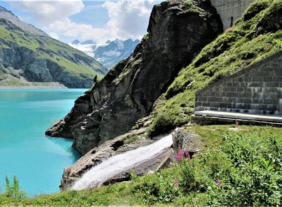 Unterhalb von Felswänden und Bergen liegt ein türkisfarbener Gletschersee, in den ein kleiner Wasserfall hinabstürzt.