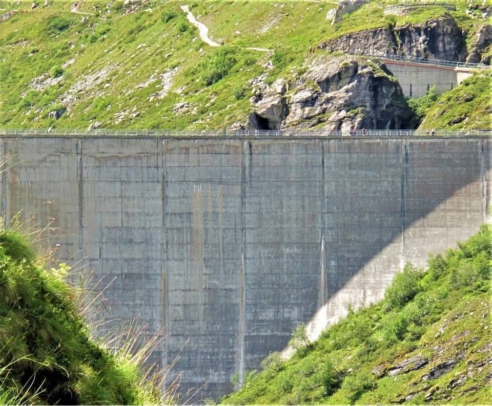 Eine massive Betongewichtsstaumauer, die ein Gebirgstal überspannt und von steilen, grasbewachsenen Hängen flankiert wird, liegt im hellen Tageslicht.