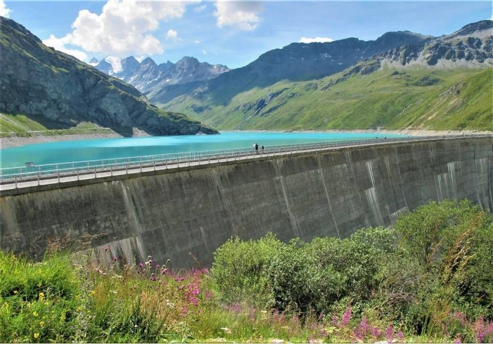 Unter einem hellen, leicht bewölkten Himmel staut ein Betonstau einen türkisfarbenen Alpensee unterhalb einer Bergkette.