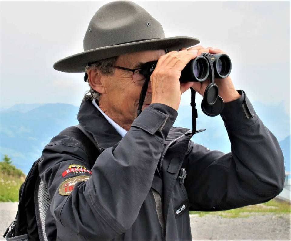Eine Person mit grauem Hut und grauer Jacke blickt durch ein Fernglas vor einer bergigen, bewölkten Kulisse.