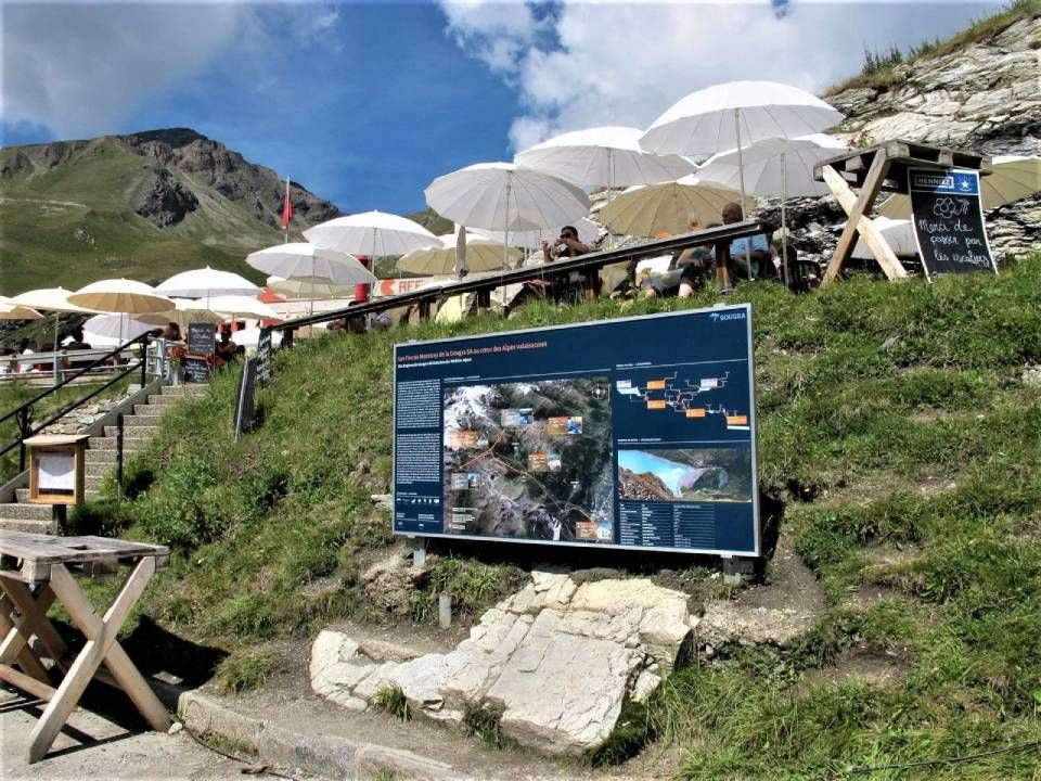 Auf einem grasbewachsenen Berghang steht eine Informationstafel in der Nähe eines Sitzbereichs im Freien mit Tischen und weissen Sonnenschirmen.