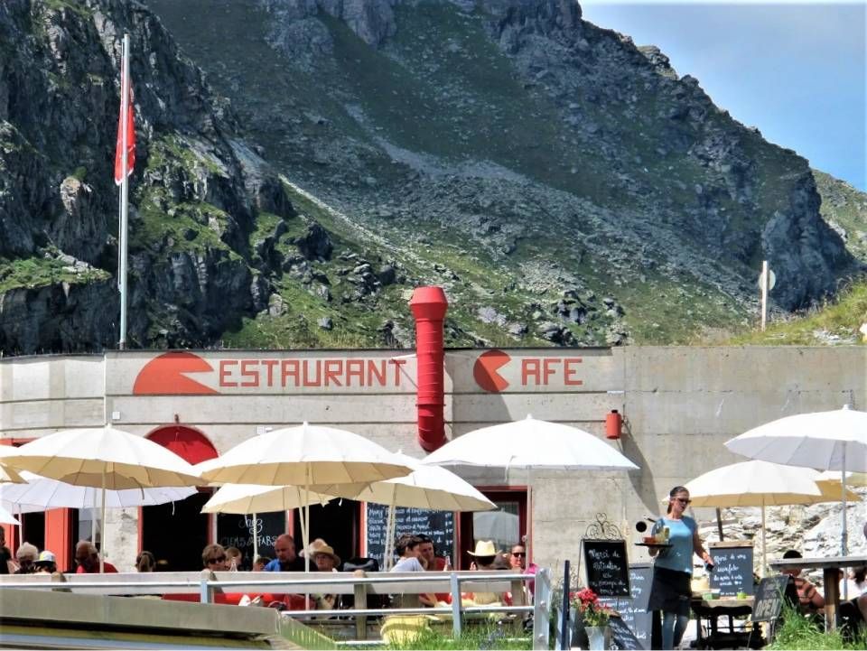 Restaurant- und Cafébereich im Freien mit weissen Sonnenschirmen am Fusse eines grossen Berges.