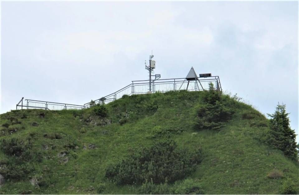 Ein grasbewachsener Berggipfel mit einer umzäunten Aussichtsplattform, einem Kommunikationsturm und einem kleinen pyramidenförmigen Dach.