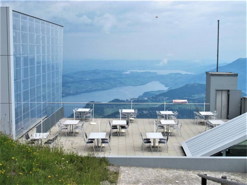 Eine Aussenterrasse mit kleinen Tischen und Stühlen mit Blick auf einen von Bergen umgebenen See an einem bewölkten Tag.