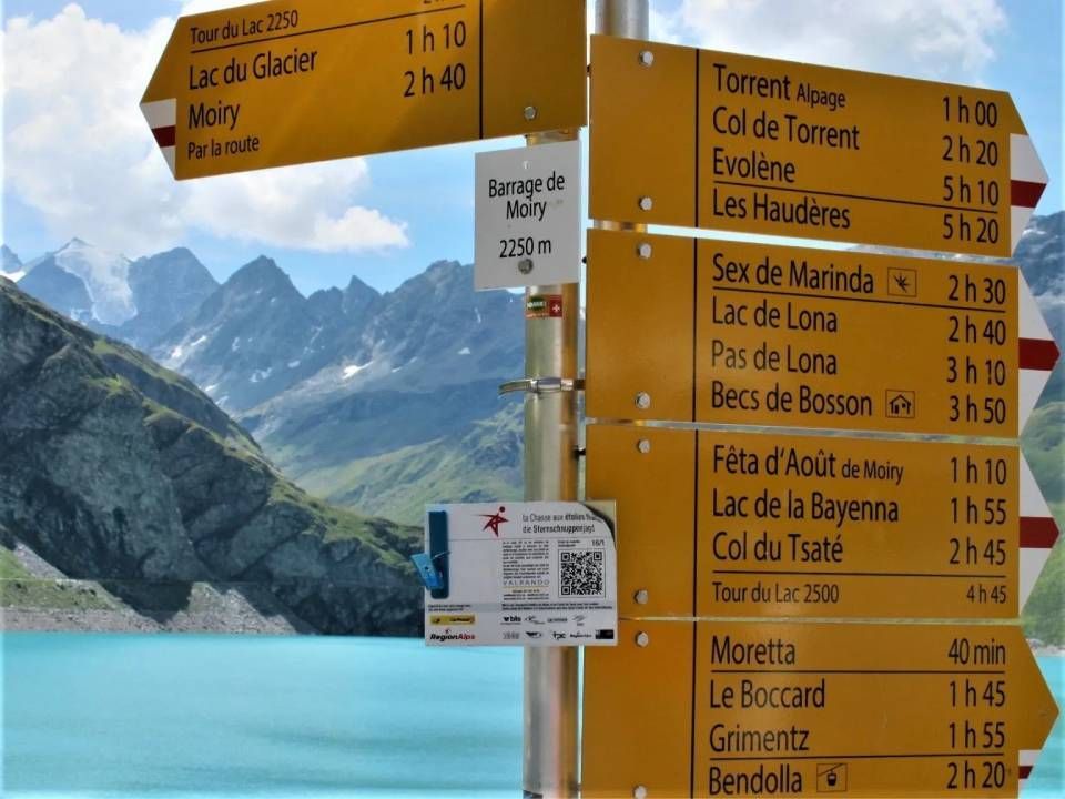 Wanderwegweiser mit Richtungspfeilen, der verschiedene Wanderwege, Gehzeiten und Ziele vor der Kulisse eines Bergsees auflistet.
