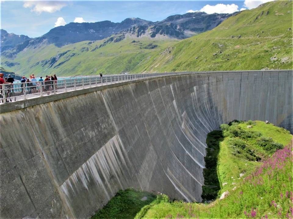 Eine massive, geschwungene Staumauer aus Beton erhebt sich aus einer grünen Berglandschaft; Menschen spazieren auf dem oberen Geländer.