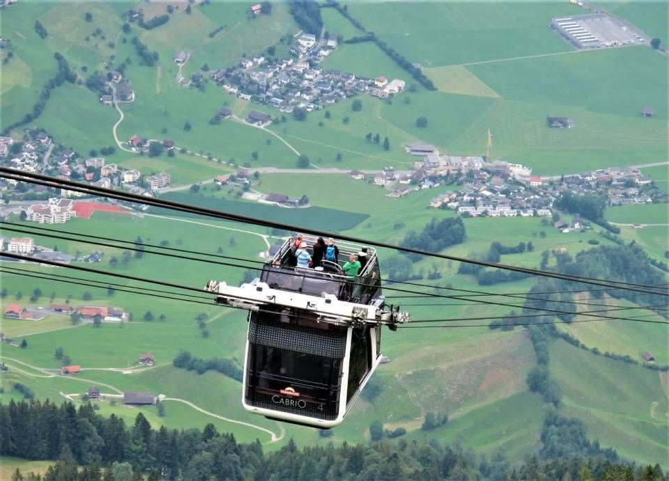 Eine doppelstöckige Seilbahn mit offenen Sitzplätzen fährt über ein üppig grünes Schweizer Tal mit kleinen Dörfern.