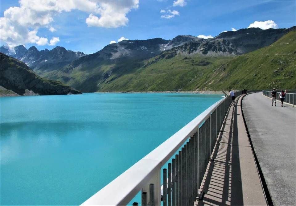 Ein malerischer Blick auf einen leuchtend türkisfarbenen Bergstausee von einem gepflasterten Dammsteg aus, mit fernen, üppigen, sanft geschwungenen Gipfeln.