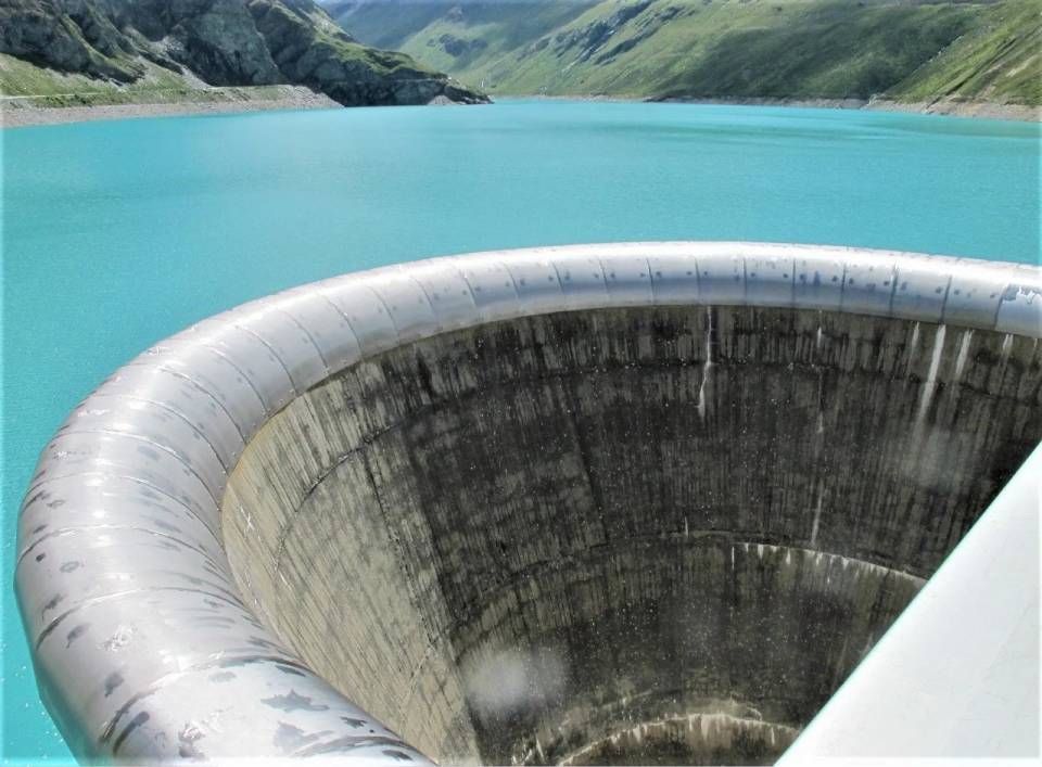 Ein grosser, kreisrunder Überlauf befindet sich in einem türkisfarbenen Stausee vor der Kulisse grüner Berghänge.
