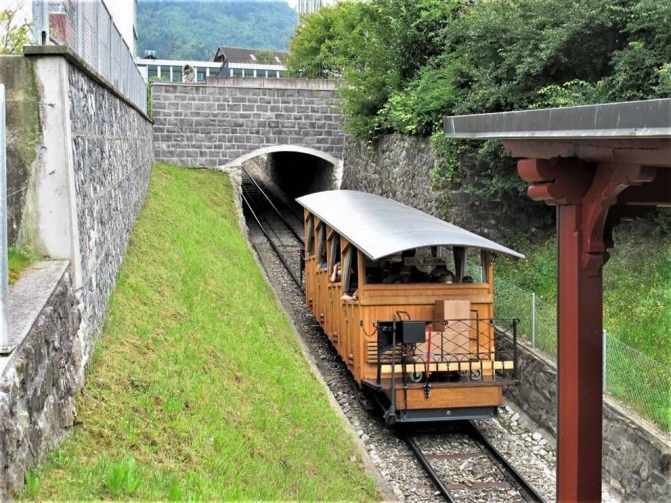 Eine hölzerne, nach beiden Seiten offene Standseilbahn-Waggon fährt in einen Steintunnel inmitten einer üppig grünen Naturlandschaft ein.