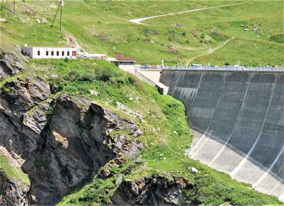 Ein grosser Betonstaudamm schmiegt sich an einen zerklüfteten, grasbewachsenen Hang; links davon steht ein kleines Gebäude.