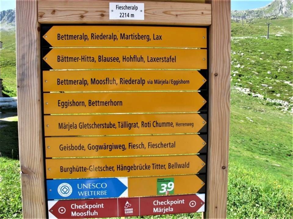 Ein hölzernes Wegweiserschild auf der Fiescheralp (2214 m) mit mehreren gelben Pfeilen, die Wanderziele auflisten, und einem UNESCO-Zeichen.